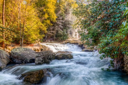 A serene river flows over rocks, surrounded by lush greenery and trees, with a gentle waterfall in the background.
