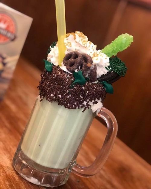 A tall glass filled with a green milkshake, topped with whipped cream, chocolate sprinkles, and various candies.