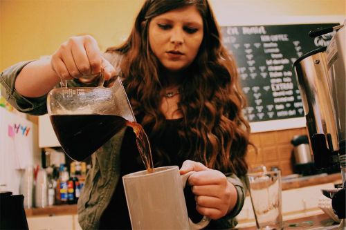 A person with long hair pours coffee from a glass carafe into a white mug in a café setting.