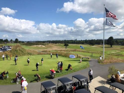 A sunny golf course scene with players practicing putting, golf carts nearby, and flags waving in the background.