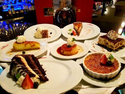 A variety of desserts displayed on white plates, including cakes, flan, and crème brûlée, with garnishes and whipped cream.
