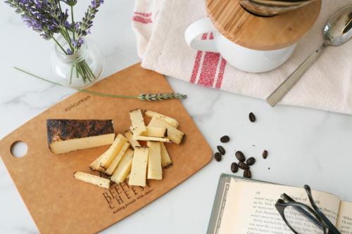 A wooden cutting board with sliced cheese, coffee beans, a book, and a vase of lavender on a marble surface.