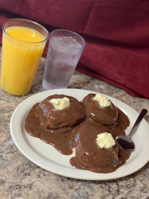 A plate of three chocolate-covered pancakes topped with butter, alongside a glass of orange juice and a glass of water.
