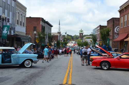 A lively street scene with classic cars on display, people gathered, and shops lining the road under a cloudy sky.