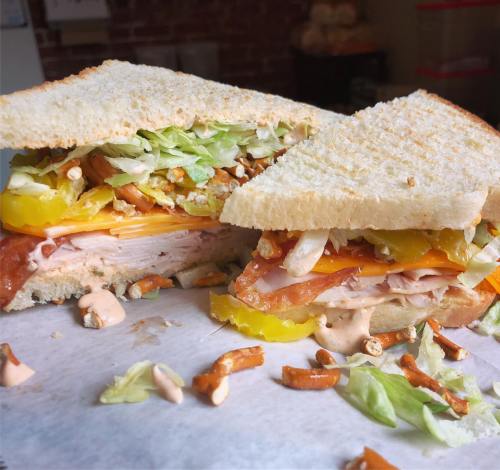 A close-up of a sandwich with turkey, cheese, lettuce, pickles, and pretzel bits, cut in half on a white surface.