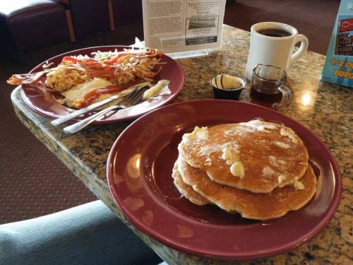 A plate of fluffy pancakes with butter, alongside a plate of bacon and hash browns, with coffee in the background.