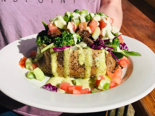 A loaded baked potato topped with fresh vegetables, drizzled with sauce, served on a white plate.