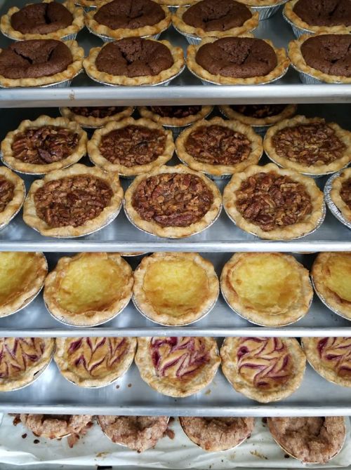 A variety of mini pies on a baking tray, including pecan, chocolate, lemon, and fruit-filled options.