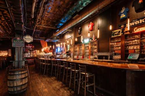 A cozy bar interior with wooden stools, shelves of drinks, and warm lighting, featuring sports memorabilia on the walls.