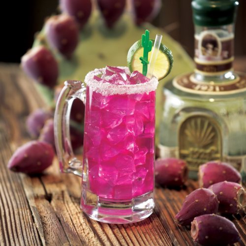 A vibrant pink cocktail with ice, garnished with a lime and cactus straw, beside a bottle and prickly pear fruit.