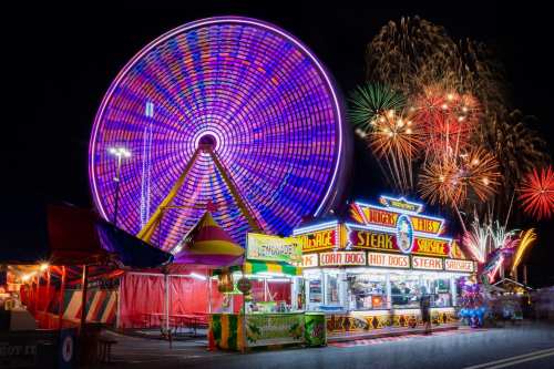 A vibrant carnival scene at night featuring a Ferris wheel, food stalls, and colorful fireworks in the background.