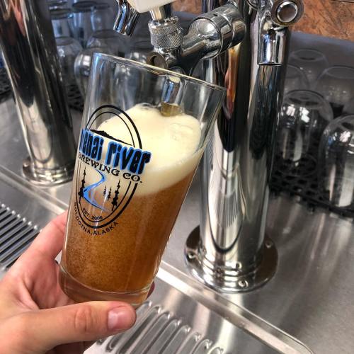 A hand holds a glass of beer being poured from a tap at a brewery, with the logo of a brewing company visible.