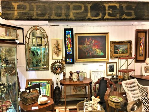 A vintage shop interior featuring various antiques, artwork, and a large sign reading "PEOPLES" above the displays.