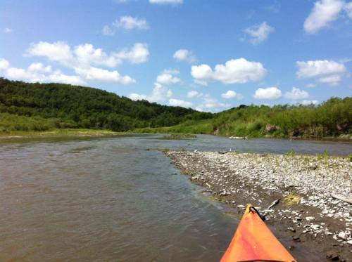 A view from a kayak on a river, surrounded by green hills and a blue sky with fluffy clouds.