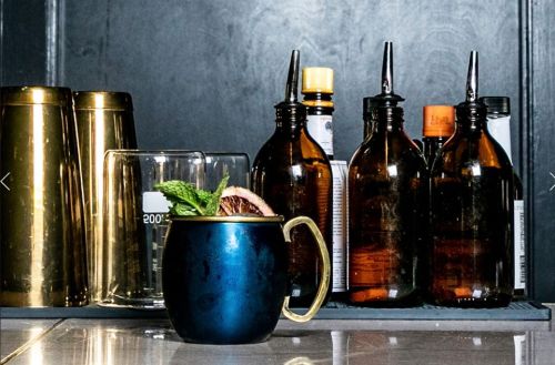 A blue mug with a garnish sits in front of various glass bottles and cocktail shakers on a bar counter.