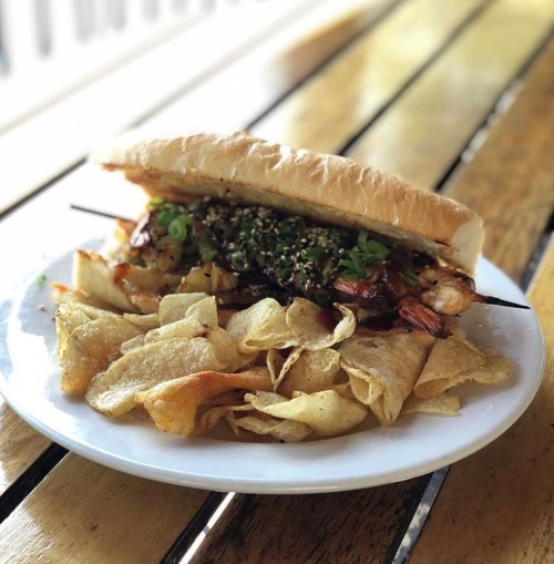 A sandwich filled with grilled shrimp and toppings, served with a side of crispy potato chips on a wooden table.