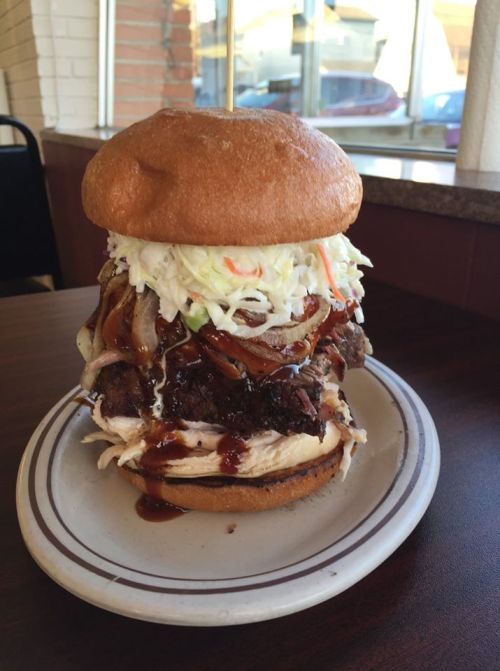 A towering burger with layers of meat, coleslaw, and barbecue sauce, served on a plate in a casual dining setting.