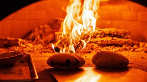 Two calzones baking in a wood-fired oven, with flames and glowing embers in the background.
