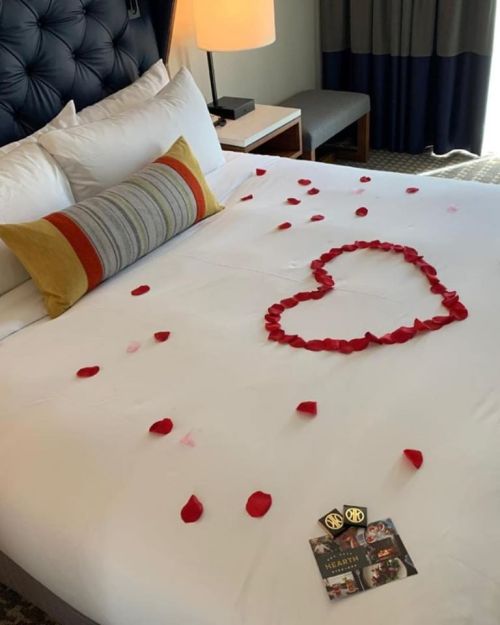 A bed with white sheets decorated with rose petals arranged in a heart shape, alongside a decorative pillow and a card.
