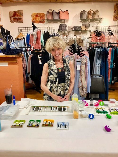 An elderly woman stands behind a table with craft supplies in a boutique, surrounded by clothing and accessories.
