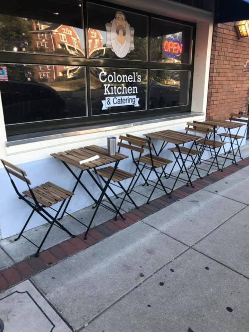 Outdoor seating with wooden tables and chairs in front of Colonel's Kitchen & Catering, with an "Open" sign in the window.