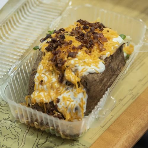 A loaded baked potato topped with cheese, bacon bits, and sour cream in a clear takeout container.