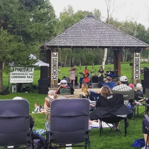A live music performance on an outdoor stage with an audience seated on lawn chairs in a park setting.