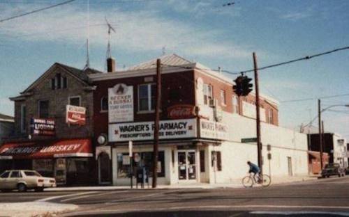 A street view of a pharmacy and nearby buildings, with a cyclist passing by and a clear blue sky above.