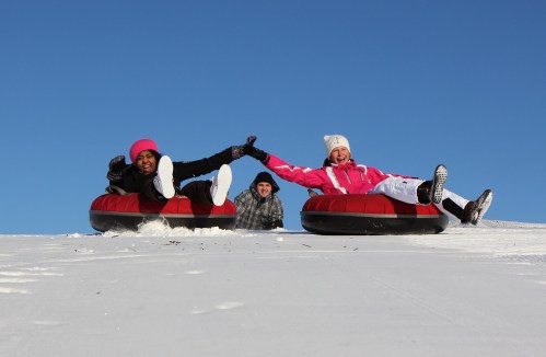 Two people in colorful winter gear joyfully sled down a snowy hill, while a third person smiles in the background.