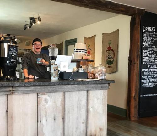 A barista smiles behind a wooden counter, preparing drinks in a cozy café with a chalkboard menu in the background.
