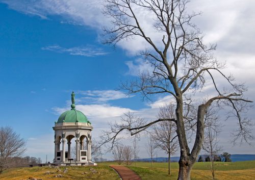 A green-domed gazebo stands on a grassy hill, with a bare tree nearby and a blue sky with clouds in the background.