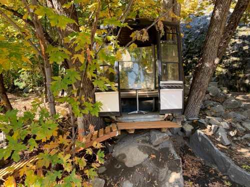 A small cabin nestled between trees, with a wooden deck and surrounded by autumn foliage and rocks.