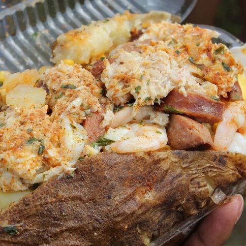 A close-up of a loaded baked potato topped with shrimp, sausage, and seasoned crab meat, garnished with herbs.
