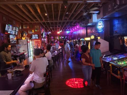 A lively arcade bar with neon lights, people socializing, and pinball machines in a vibrant atmosphere.