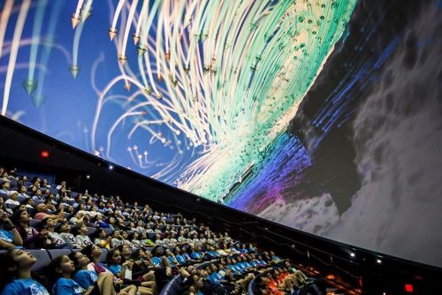A large audience watches a colorful, dynamic projection on a dome screen in a theater.