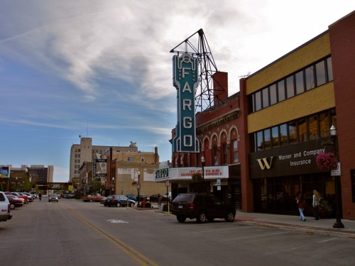 Historic downtown Fargo with a vintage theater sign, shops, and parked cars under a blue sky.