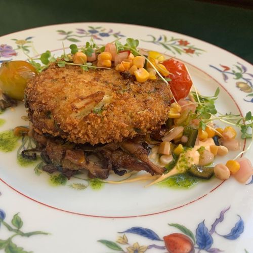 A beautifully plated dish featuring a crispy patty, garnished with corn, cherry tomatoes, and microgreens on a decorative plate.