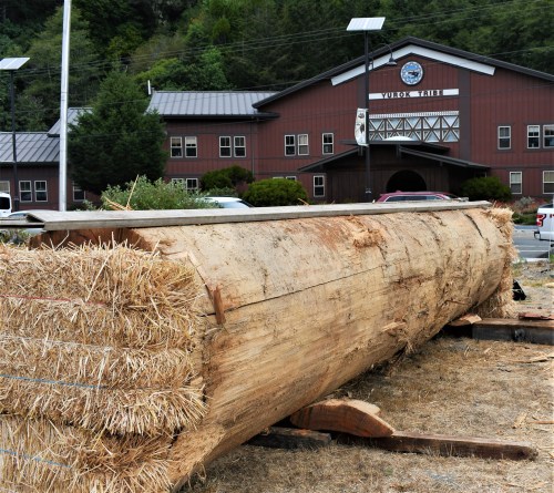 A large log rests on straw bales in front of a building with a sign reading "Vulcan Table" and surrounding greenery.