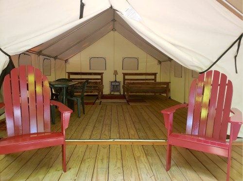 Interior of a glamping tent featuring two red Adirondack chairs, a table, and two wooden beds on a wooden floor.