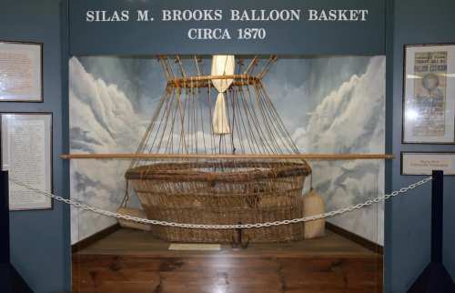 Display of Silas M. Brooks' balloon basket from circa 1870, surrounded by historical documents and a cloud backdrop.