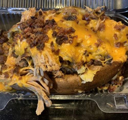 A loaded baked potato topped with shredded meat, melted cheese, and crispy bacon bits in a takeout container.