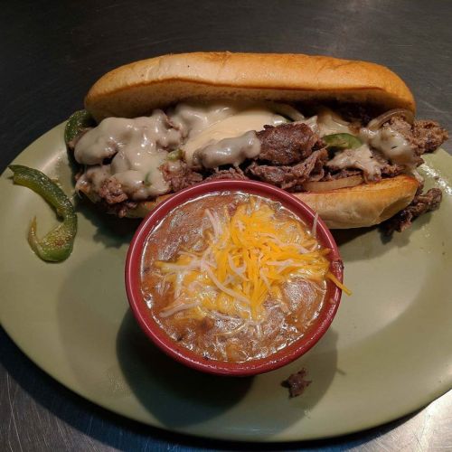 A plate with a cheesy beef sandwich topped with peppers and a bowl of chili garnished with shredded cheese.