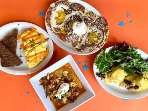 A colorful spread of brunch dishes: pancakes, omelette, salad, and a seafood stew on an orange table.