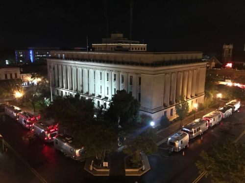 Aerial view of a large building at night, surrounded by several illuminated vehicles parked along the street.