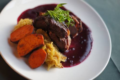 A plate of sliced meat with a rich sauce, served with sweet potatoes and sauerkraut, garnished with green onions.