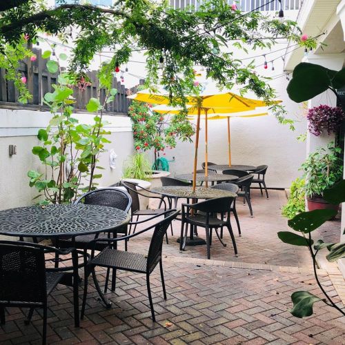 A cozy outdoor patio with black tables and chairs, yellow umbrellas, and lush greenery.