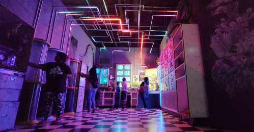 A vibrant room with neon lights and a checkered floor, featuring people interacting with colorful displays.