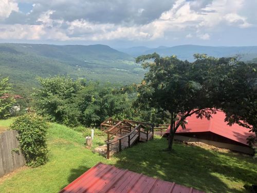 A scenic view of rolling green hills and mountains under a partly cloudy sky, with trees and a red-roofed building in the foreground.