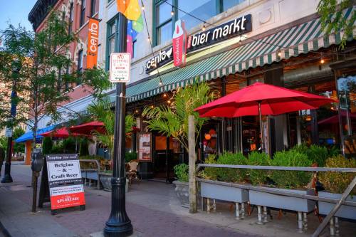 A lively street scene featuring a bar with red umbrellas, outdoor seating, and a sign promoting Belgian craft beer.