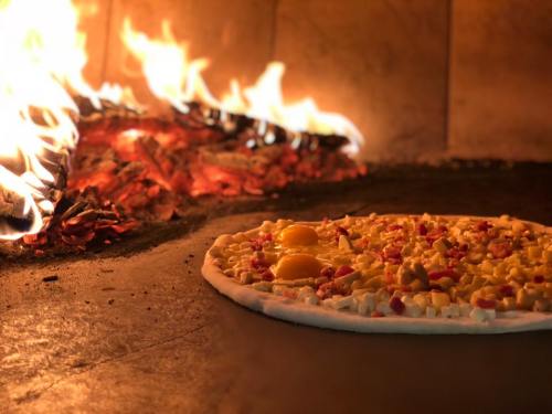 A pizza with toppings and raw eggs baking in a wood-fired oven, with flames in the background.